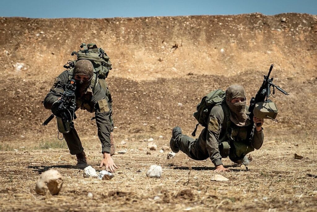 Two special forces operatives in tactical gear demonstrating low-crawl movement in a desert-like environment.