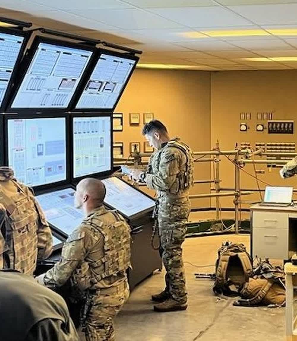 Military personnel in tactical gear monitor complex systems in what appears to be a nuclear facility control room or training simulator.