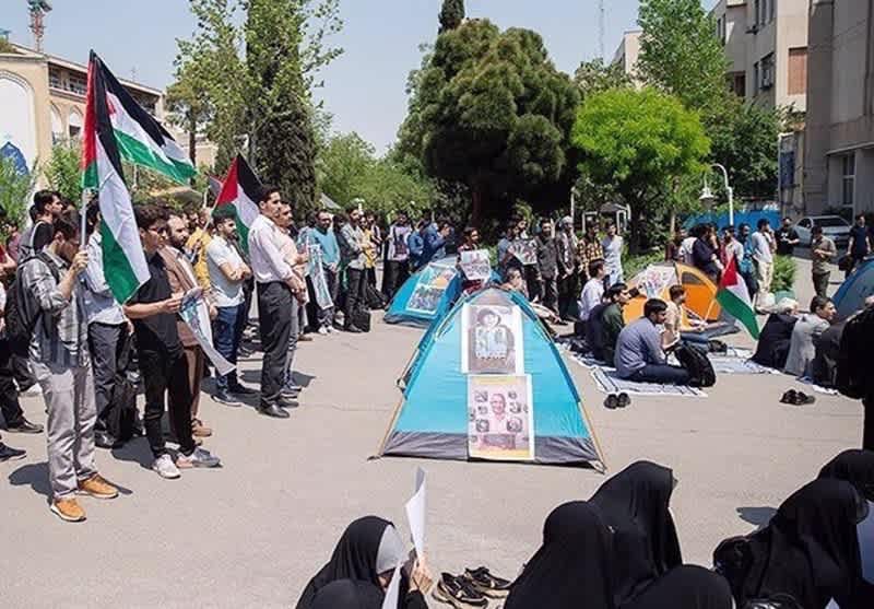 Iranian students gather outdoors for a rally, holding Palestinian flags and seated around tents with displayed posters.