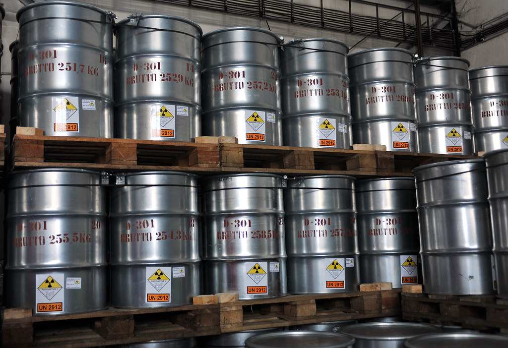 Metal drums containing yellowcake uranium powder, marked with radioactive hazard symbols, are stored on wooden pallets.
